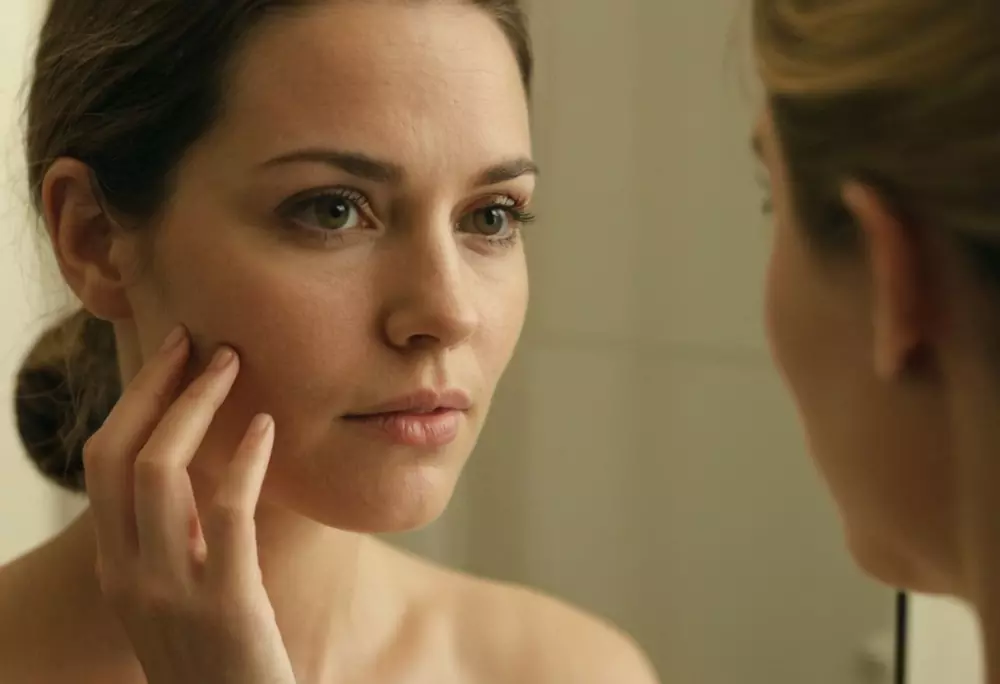 Nahaufnahme einer Frau, die ihr Gesicht in einem Spiegel untersucht, wobei der Fokus auf den Hautveränderungen nach der Schwangerschaft liegt. Aufgenommen in einem gut beleuchteten Badezimmer, was eine nachdenkliche Stimmung widerspiegelt. Fotografisch, mit Makro-Objektiv, um die detaillierte Hautstruktur einzufangen, wobei natürliche Beleuchtung und hohe Auflösung betont werden.
