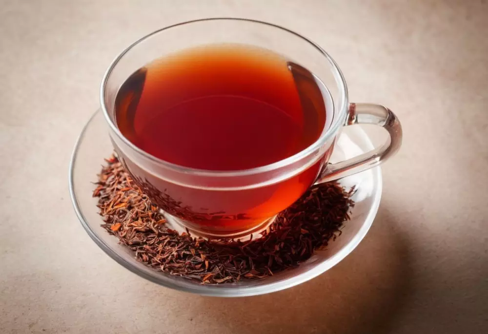Hojas de té Rooibos rojo intenso y una taza clara de té preparado, presentados en un fondo minimalista para resaltar el vibrante color rojo. Estilo fotográfico con un enfoque en el contraste de colores y la iluminación natural.
