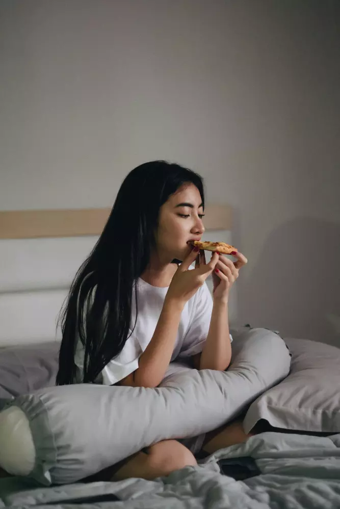 Eine Frau sitzt auf einem Bett und isst ein Stück Pizza.