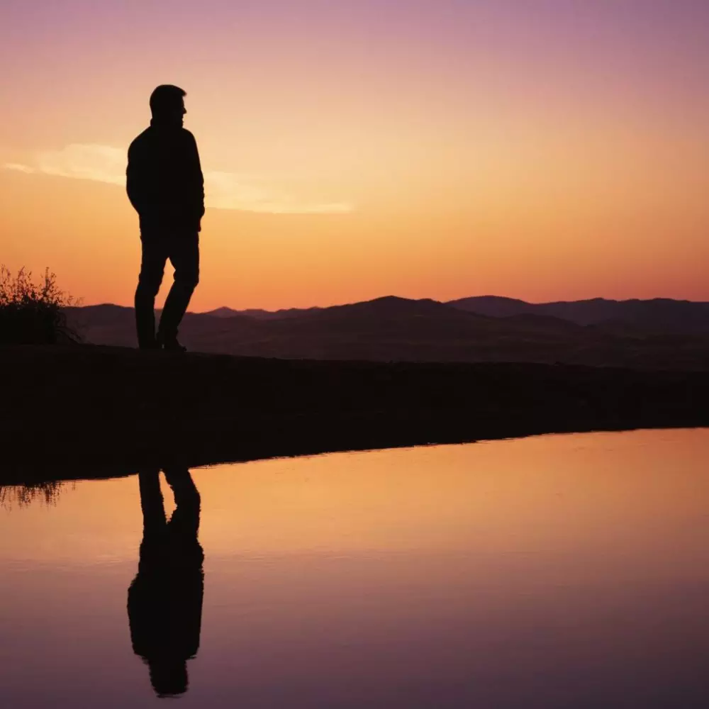 Solitary man silhouette against a sunset, looking into the distance, depicting reflection and introspection, in a cinematic style.