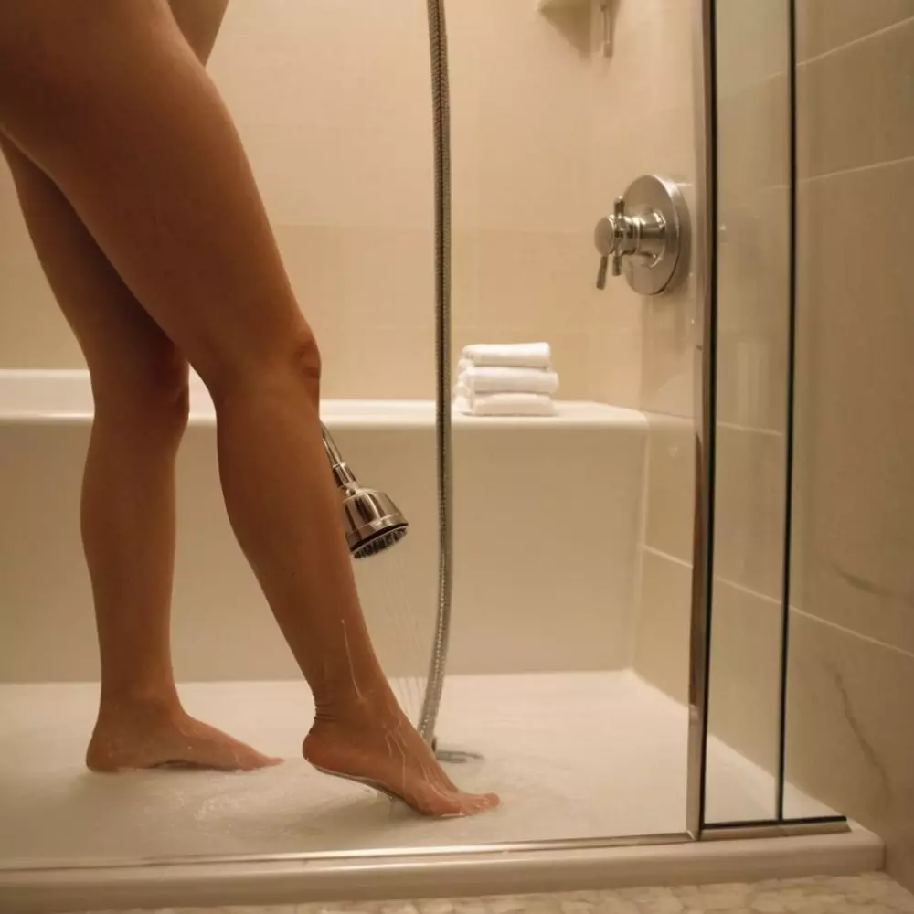 Person benutzt eine Handbrause an ihren Beinen in einem Badezimmer, um den Prozess des Wechsels zwischen heißem und kaltem Wasser zu demonstrieren. Das Foto wurde mit weichem Badezimmerlicht und einem mittleren Teleobjektiv aufgenommen.